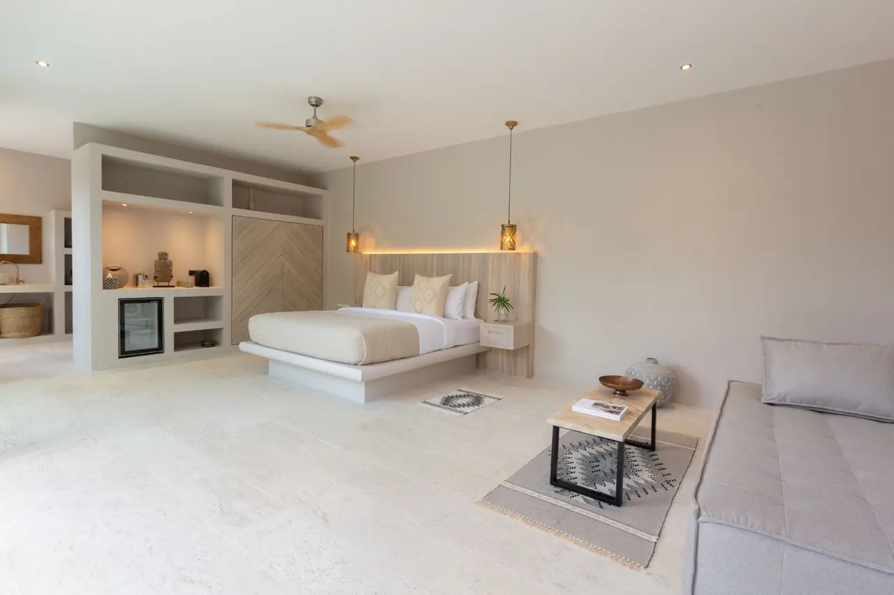 Wide, bright bedroom with a platform bed, lantern pendants, a ceiling fan, built-in shelves with a minibar, and a coffee table and daybed in the foreground.