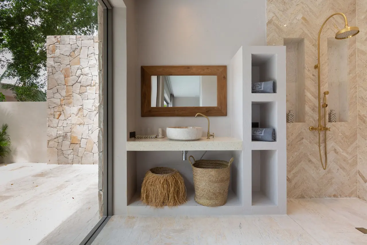 Bathroom with a stone basin under a teak-framed mirror, woven baskets, Cielo towels in wall niches and a brass rain shower on herringbone tiles, open to a stone-walled garden.