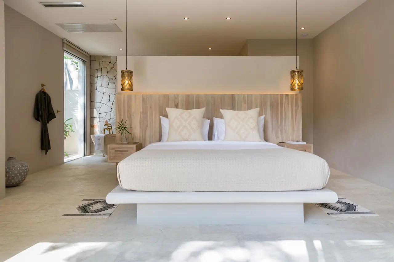 Bed on a white plinth against a timber headboard between two brass lantern pendants, with a stone-walled bathroom and a robe on a hook at left.
