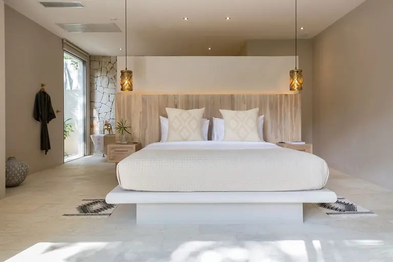 Bed on a white plinth against a timber headboard between two brass lantern pendants, with a stone-walled bathroom and a robe on a hook at left.