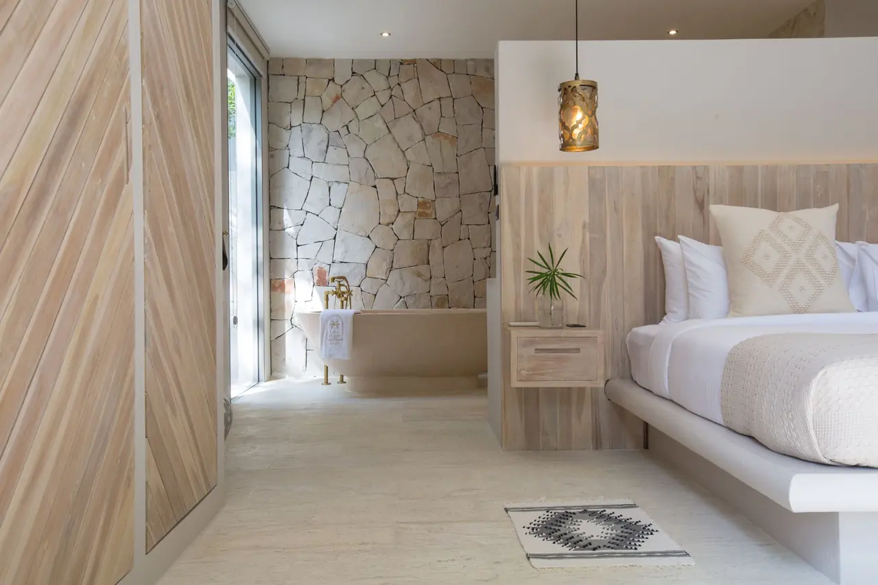 Open-plan suite with chevron timber doors, a freestanding stone bathtub against a rough stone wall and the bed beside a timber headboard.