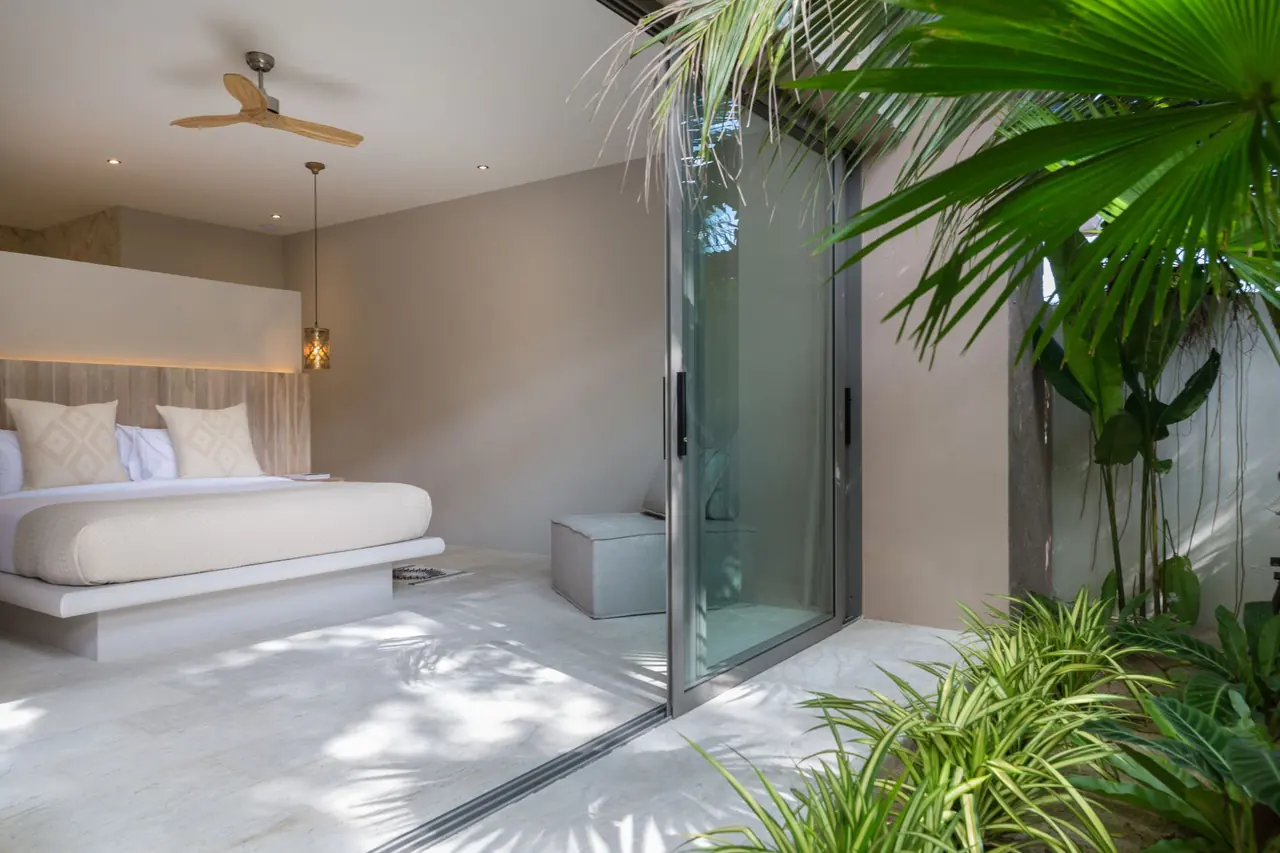 Bedroom with a ceiling fan and a platform bed, its glass sliding door open to a planted garden in dappled sunlight.