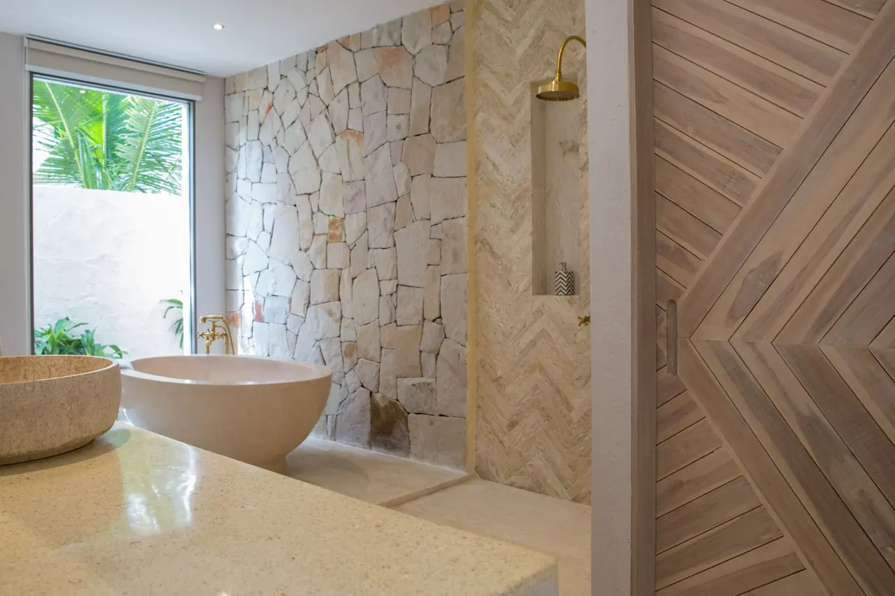 Bathroom with a stone basin on a terrazzo counter, a freestanding bathtub, a stone wall, a brass rain shower on herringbone tiles and a window onto palms.