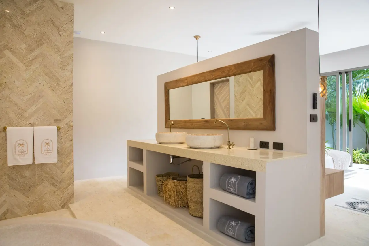 Double vanity with two stone basins under a teak-framed mirror, Cielo logo towels, woven baskets and a view through to the bedroom and garden.