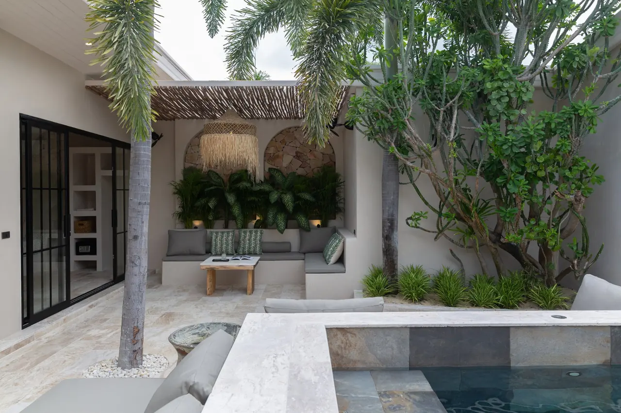 Villa courtyard with a palm and a leafy tree, a raised slate-lined pool and a covered lounge with stone arches and a raffia pendant, beside black-framed glass doors.