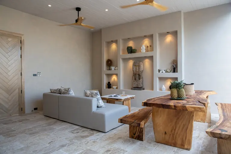 Open-plan living room with a grey sofa, a wall of lit niches holding a carved wooden figure and ceramics, and a live-edge dining table with benches and pineapples.