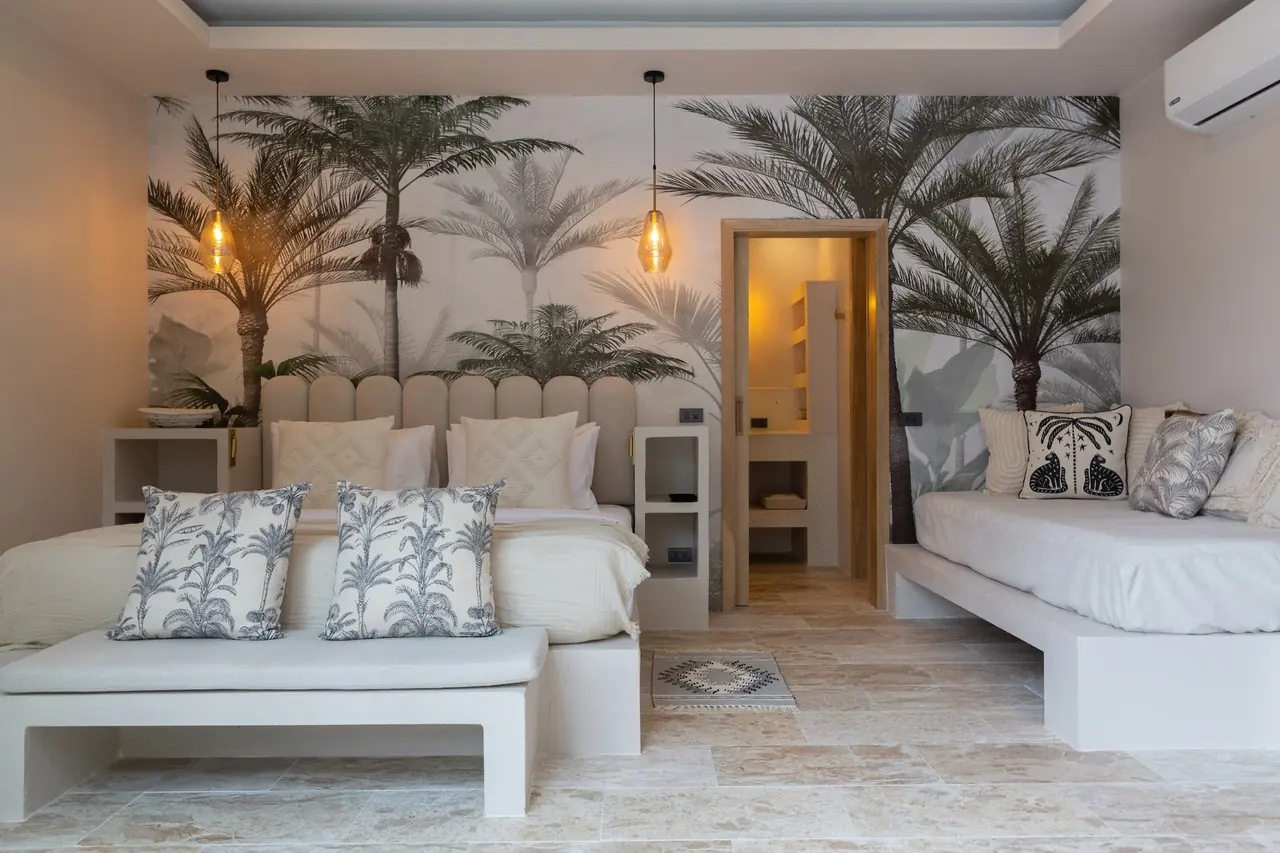 Bedroom with a palm-tree mural wall, a bed with an upholstered headboard and palm-print cushions on a bench, a daybed at right and a lit doorway to the bathroom.