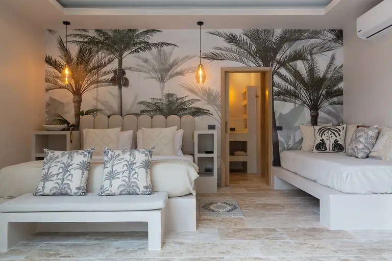Bedroom with a palm-tree mural wall, a bed with an upholstered headboard and palm-print cushions on a bench, a daybed at right and a lit doorway to the bathroom.