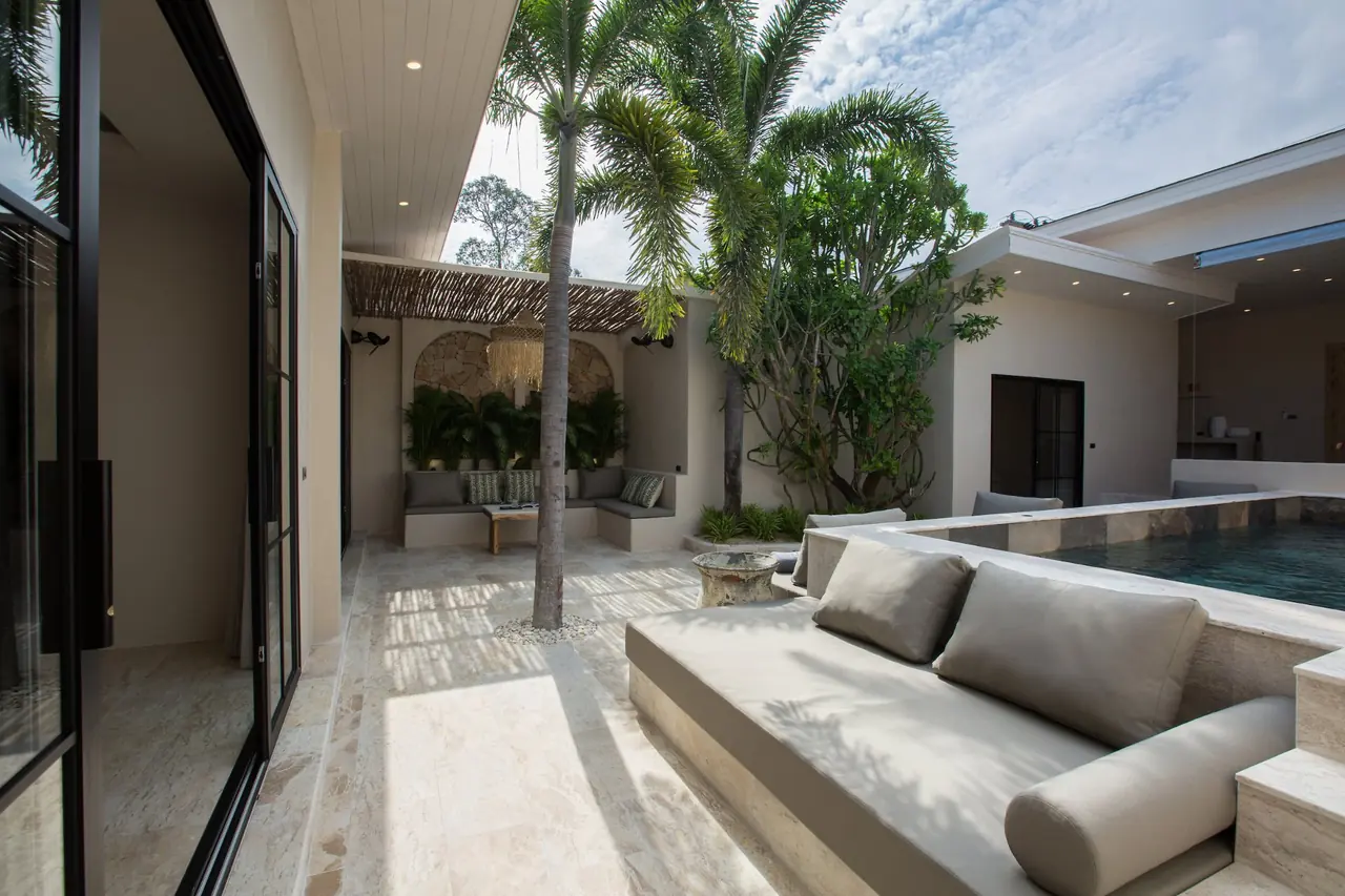 Built-in daybed with grey bolster cushions beside the raised pool, with palms and the covered pergola lounge beyond.